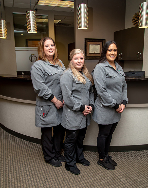 Three individuals standing in a reception area, all wearing professional attire with name tags. The person on the left is seated at a desk with a computer monitor and keyboard, while the other two stand behind them.