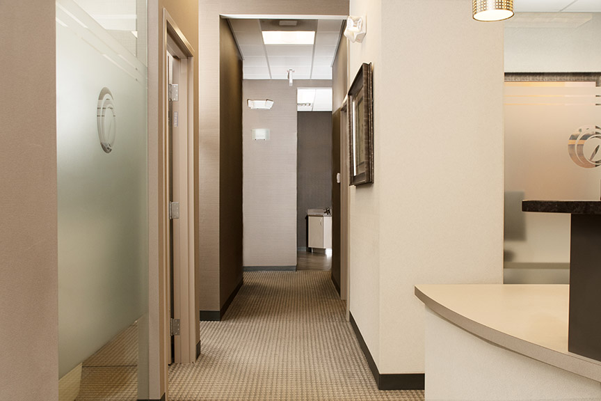 The image shows an interior space that appears to be a professional office or business environment, featuring a hallway with a modern design, including tiled flooring, walls with a combination of glass and solid panels, and a sink area at the end with a countertop.