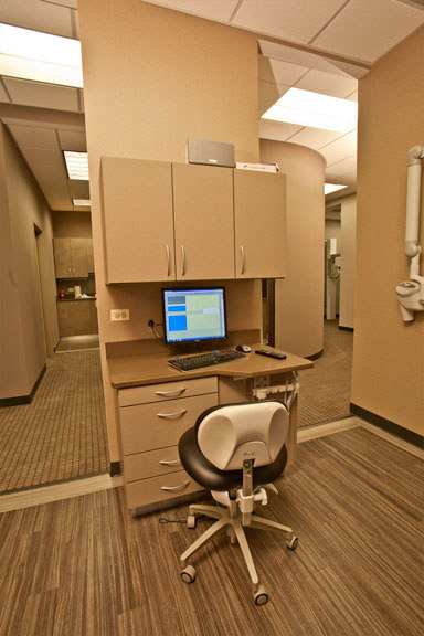 The image shows an interior space that appears to be a medical or dental office, featuring a reception area with a counter and computer monitor, a waiting room with chairs, and an open door leading into another room.