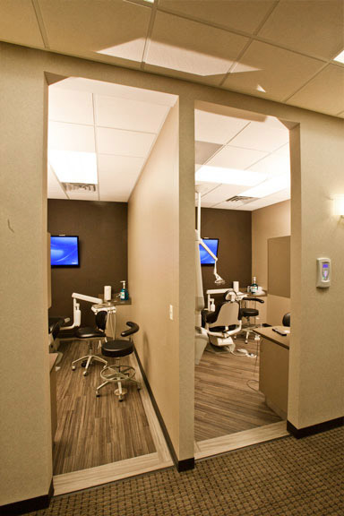 The image shows an interior view of a dental office, featuring a reception area with a desk and chairs, a waiting room with a television, and a hallway leading to the treatment rooms.