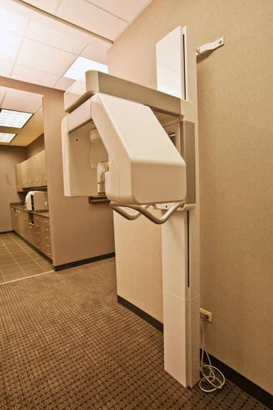 An image of a modern dental office with a large, white dental chair and a mounted digital X-ray machine, situated in front of a beige wall in an interior space.