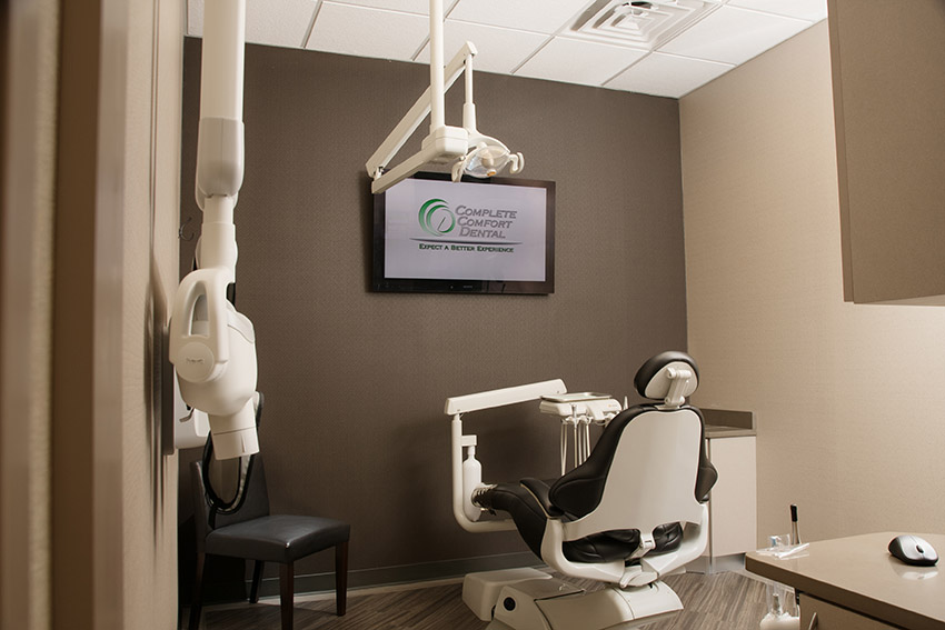 The image shows a modern dental office interior with a dental chair, overhead lights, and equipment, including a computer monitor displaying text.