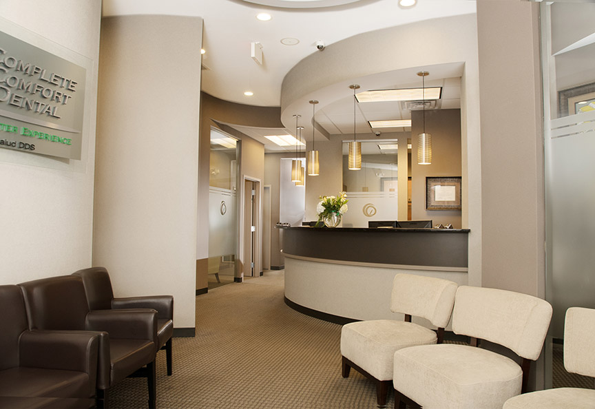 The image shows an interior space of a modern office or waiting area, featuring a reception desk, seating area with chairs and ottomans, a sign with the name  Comfort Dental , and a contemporary design with neutral colors.