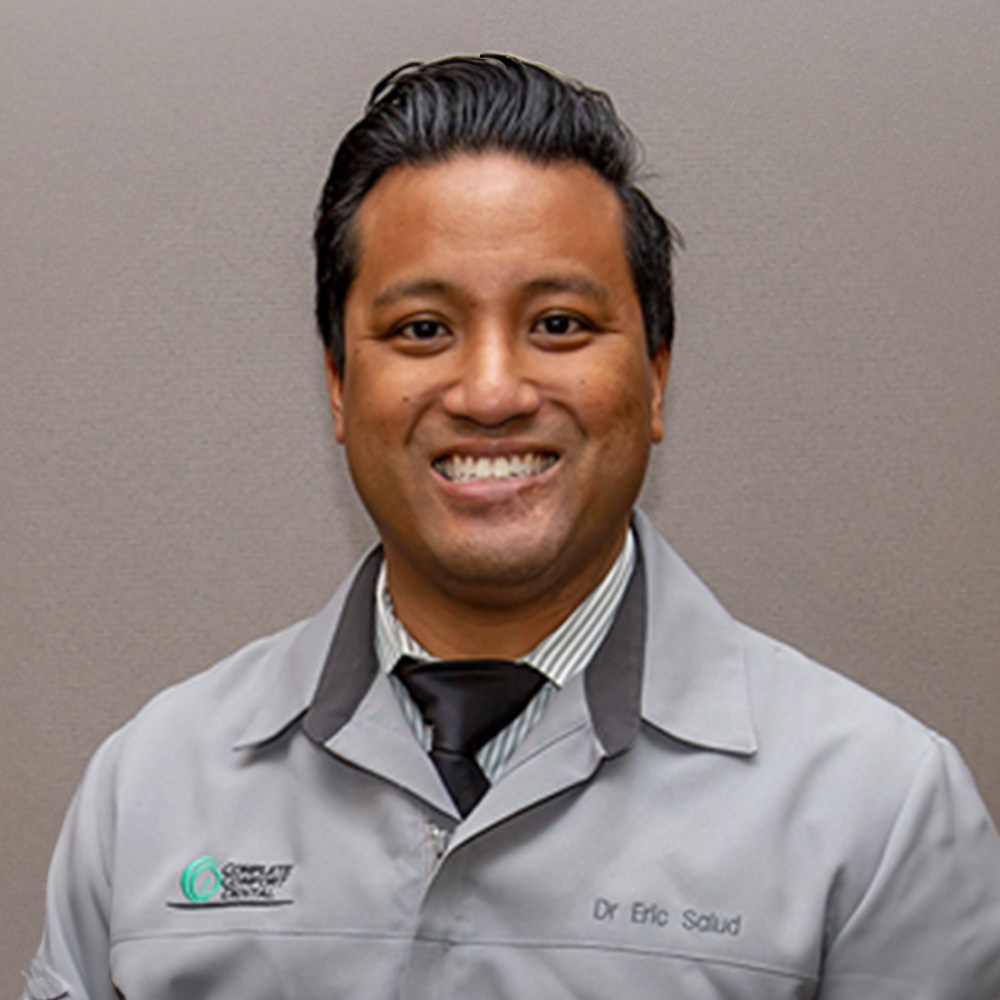 The image shows a person posing for the camera, smiling and wearing a white lab coat with name tags, suggesting a professional setting related to science or medicine.