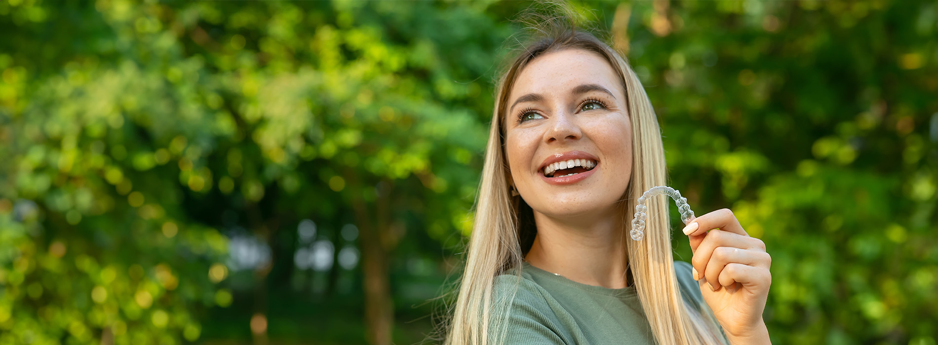 A smiling woman with blonde hair, wearing a green top, holding a key and standing in front of trees.