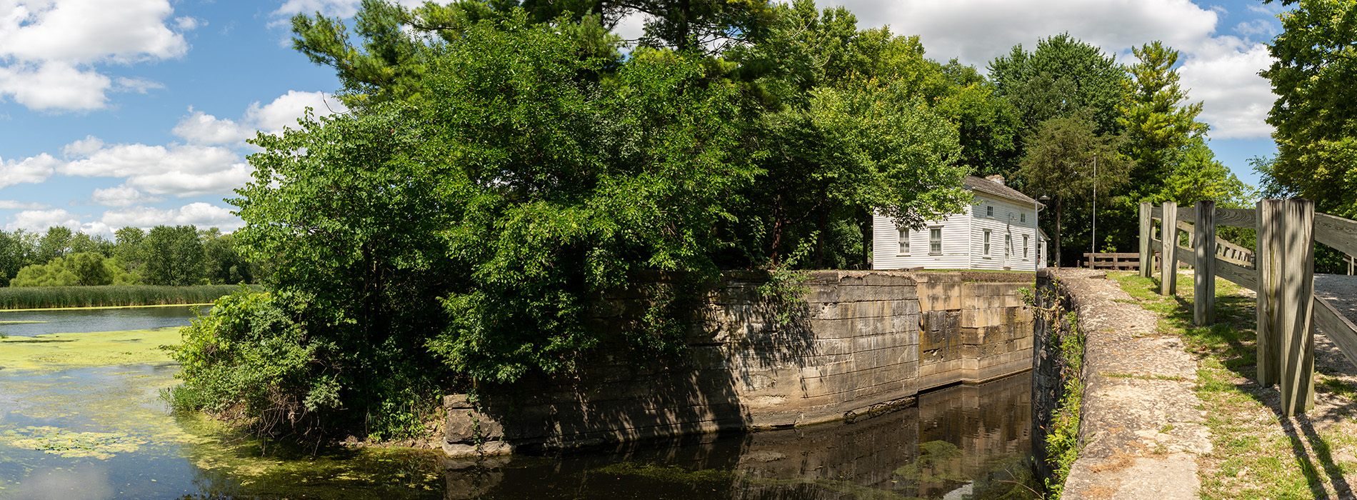 The image shows a serene outdoor scene with a river, a bridge, a small structure, and lush green trees.