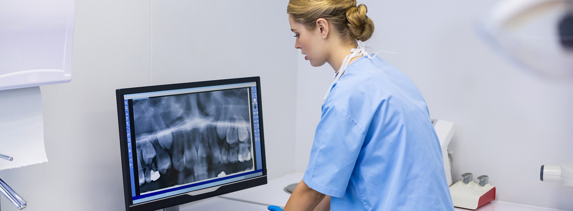 A woman in a blue uniform, standing at a medical workstation with a digital X-ray monitor.