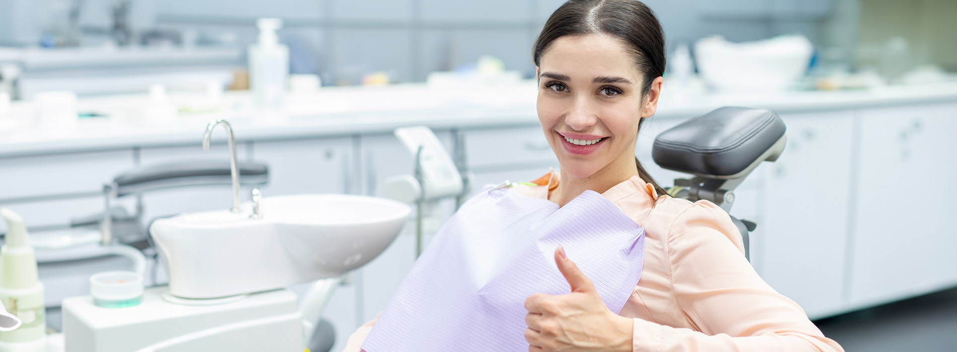 The image shows a woman in a dental office, smiling and giving a thumbs-up sign while seated on a dental chair.