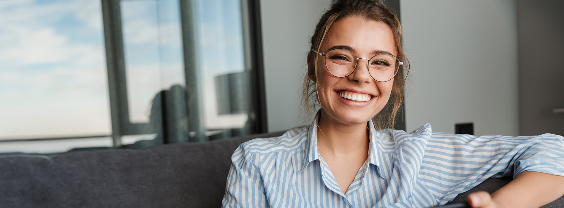 A woman with a smile, wearing glasses and a striped shirt, sits on a couch against a blurred background.