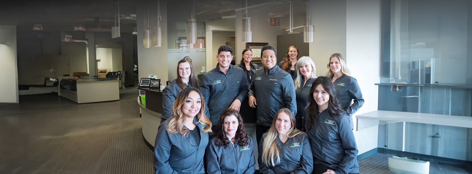 The image is a photograph of a group of individuals, likely professionals, posing together in an office environment. They are standing in front of a modern reception desk with a sleek design, and behind them is a well-lit space featuring contemporary decor.